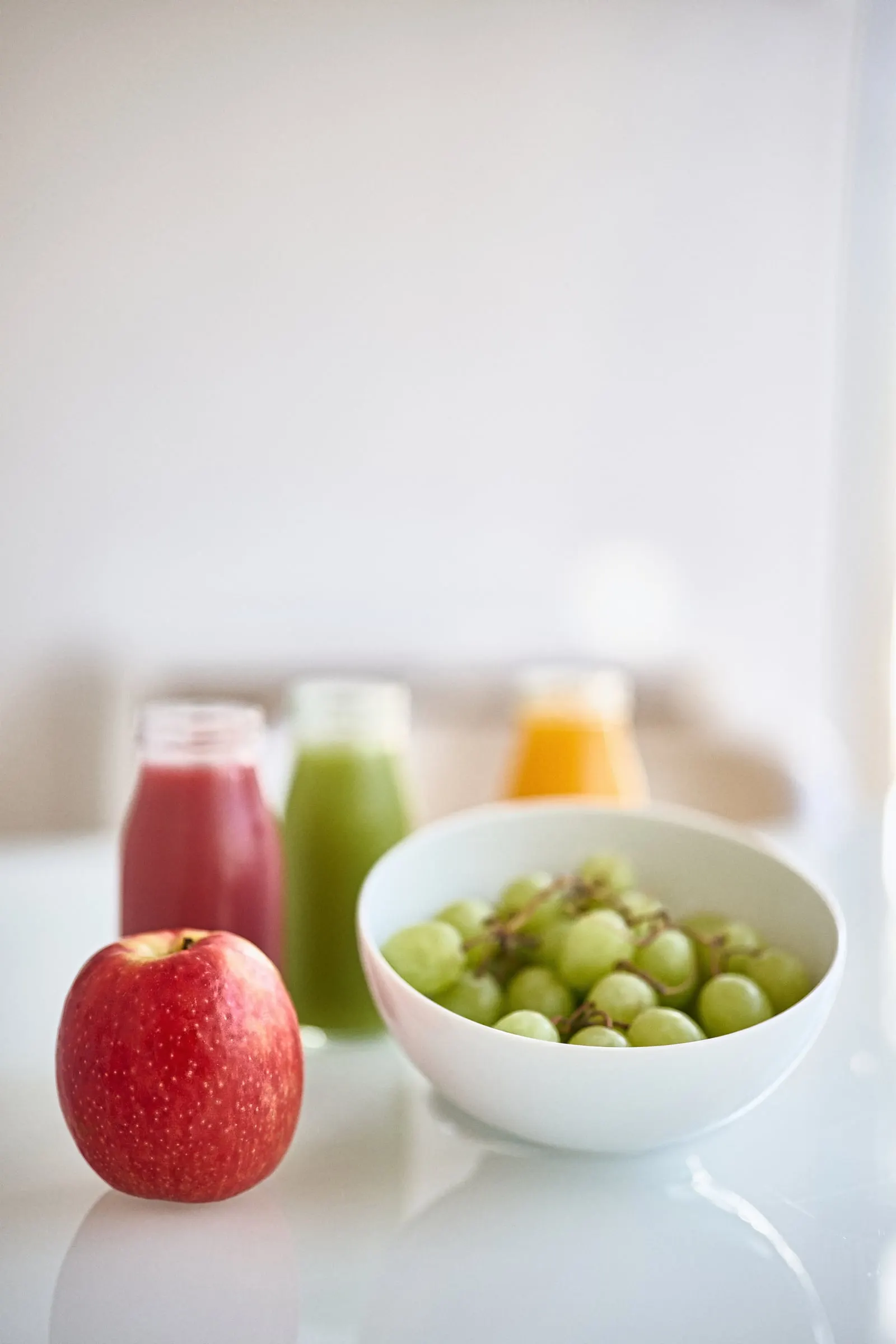 Uma maçã, uma taça de uvas e três sumos, numa mesa branca.