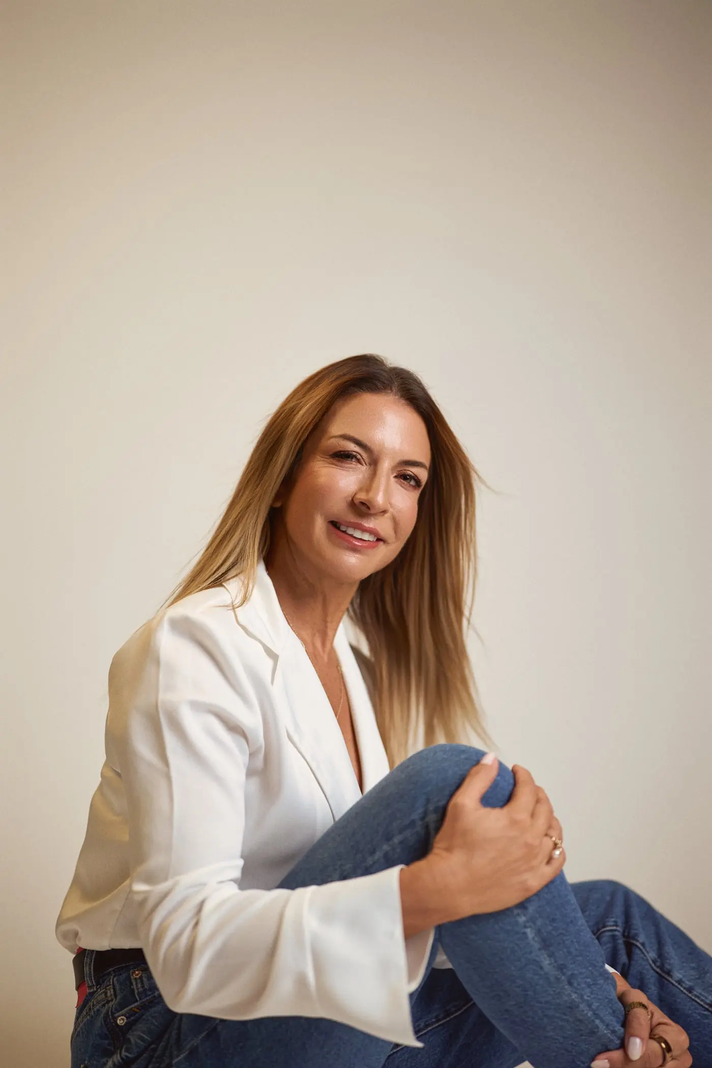 Sara Torres sentada no chão, a sorrir, de casaco branco, a abraçar um joelho.